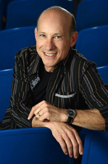 A smiling man in a black and grey striped shirt leans against blue theater seats.
