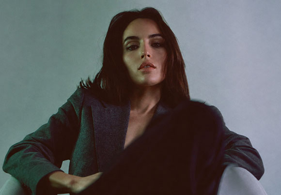 A woman with dark hair wearing a grey blazer, looking forward with a neutral expression.