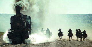 A black steam train emitting white smoke as several people ride horses alongside it on a dusty plain.