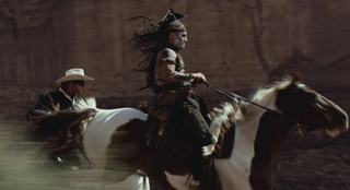 A person with dark hair and face paint rides a brown and white horse past a person in a white cowboy hat.