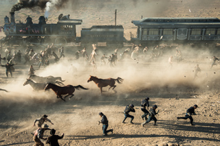 People and horses run through thick dust clouds next to a moving steam train in a desert landscape.