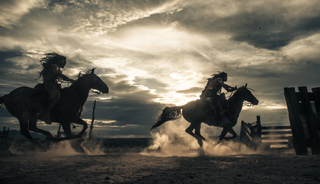 Silhouettes of two riders on galloping horses kicking up dust against a cloudy sunset sky.