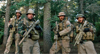 Four soldiers in tan and camouflage gear holding rifles while standing among evergreen trees.