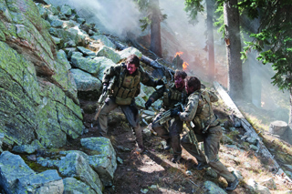 Three soldiers in camouflage gear navigate a steep, rocky, and smoky wooded slope.