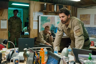 A man in tan military fatigues leans over a desk in a room filled with maps, computers, and supplies.