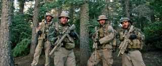 Four soldiers in desert camouflage and tactical gear holding rifles in a dense forest.