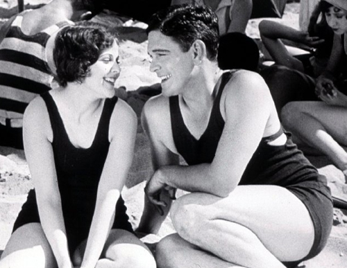 A man and woman in dark vintage one-piece swimsuits sit on a sandy beach, smiling and looking at each other.