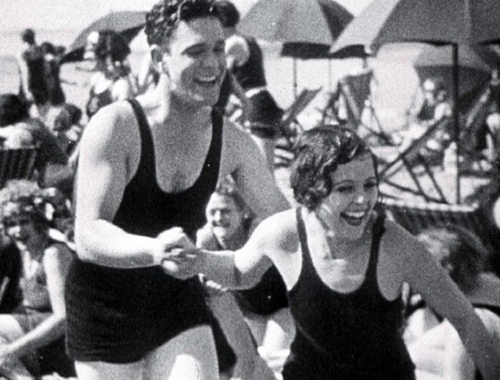 A man and woman in dark vintage swimwear laugh together on a crowded beach with umbrellas.