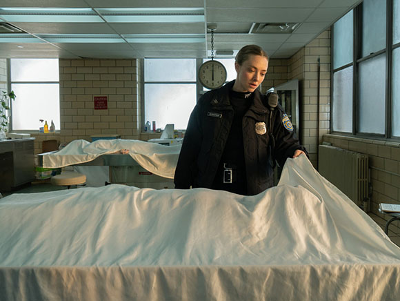 A woman in a dark uniform stands in a clinical room next to a body covered by a white sheet.