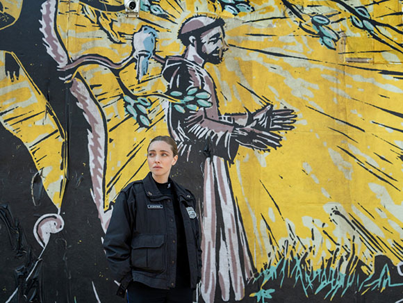 A woman in a black jacket stands before a large yellow mural depicting a man with rays of light.