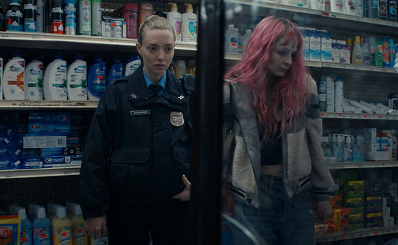 A female police officer in uniform watches a woman with long pink hair in a store aisle.