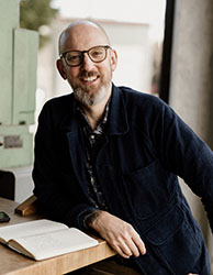 A smiling man with glasses and a graying beard wearing a dark jacket, sitting at a wooden table with an open book.