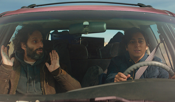 A man with a beard and a woman driving a car, both looking startled with wide eyes.