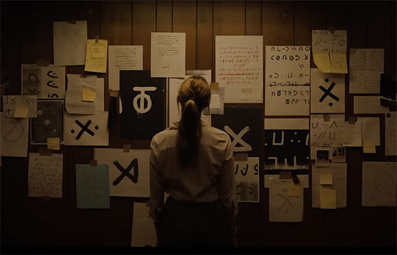 A woman viewed from behind looks at a dark wall covered in various papers, symbols, and handwritten notes.