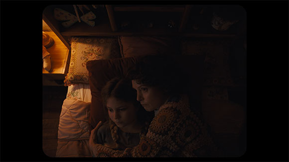 A young person and an older person lie close together in a dark bed surrounded by pillows.