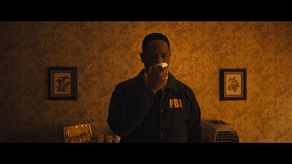 A man wearing a dark jacket with FBI printed on the chest eats food in a dimly lit room.