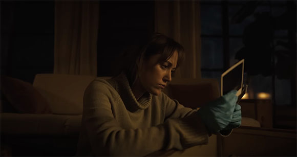 A woman in a sweater wearing a blue glove looks intently at a small rectangular device in a dark room.