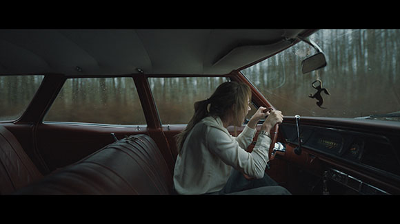 A woman in a white sweatshirt sits in the driver's seat of a vintage car on a rainy day.