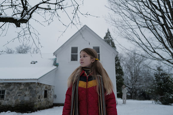 A young girl in a red quilted jacket and striped scarf stands in a snowy yard before a white house.