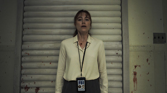 A woman in a white shirt and grey skirt stands before a shuttered door, wearing an FBI ID badge.