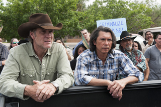 Two men, one in a cowboy hat and one in a plaid shirt, look ahead from a truck bed during a demonstration.
