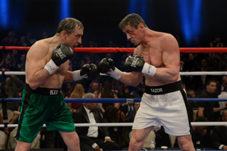 Two shirtless male boxers in a ring wearing gloves and shorts, facing each other in a combat stance.