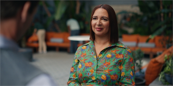 A woman with shoulder-length brown hair wearing a green floral shirt looks toward a man in a blurred foreground.