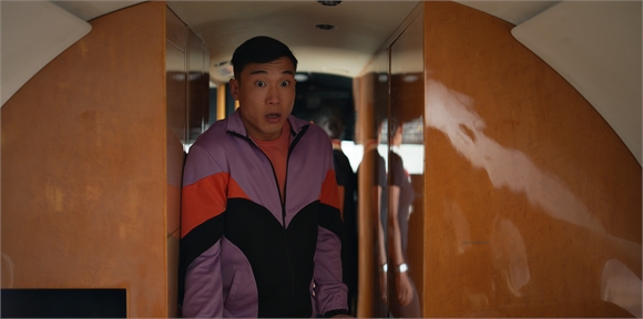 A man in a purple and orange jacket looks ahead with a wide-eyed, surprised expression in a narrow corridor.