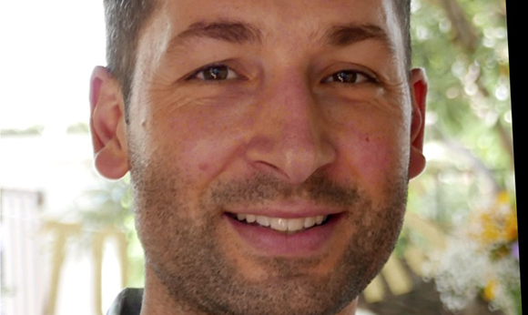 Close-up of a man with short brown hair and stubble smiling at the camera.