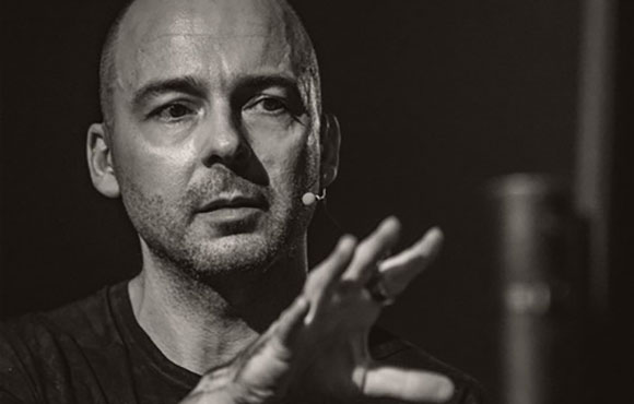 Black and white portrait of a bald man with stubble gesturing with his hand while speaking.