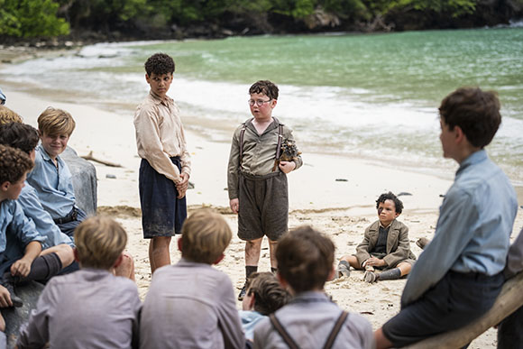 A group of boys in vintage clothing gathered on a sandy beach near the ocean.