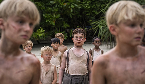 A group of young boys standing on a sandy beach surrounded by green foliage.