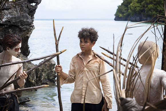 A person in a tan shirt holds a stick while two others with white body paint stand on a tropical beach.