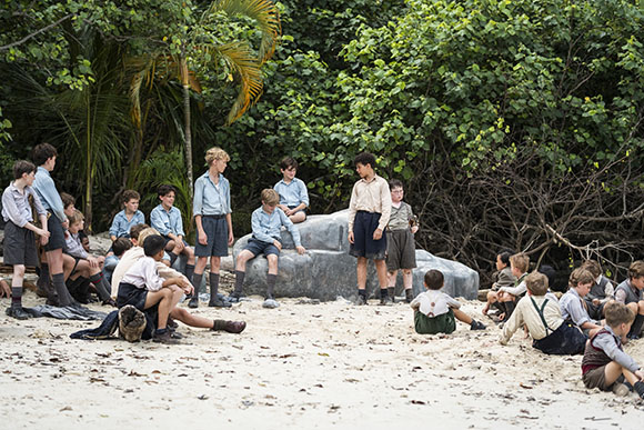 A group of boys in vintage school uniforms sitting and standing on a sandy beach near dense green trees.