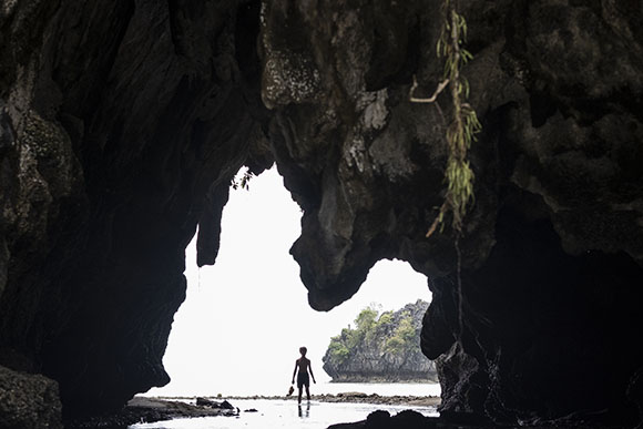 Silhouette of a person standing in a dark cave opening facing a calm ocean and rocky islands.