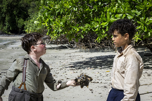 A boy in suspenders holds out a large conch shell toward another boy standing on a sandy beach.