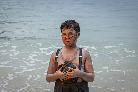A young boy with glasses and dirt on his face holds a large seashell while standing on a sandy beach.
