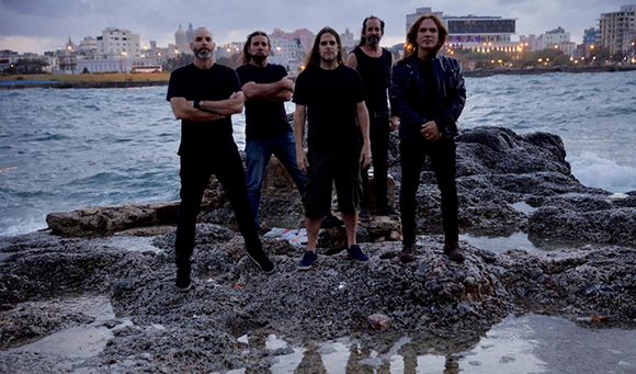 Five men in dark clothing standing on jagged rocks by the ocean under a cloudy sky.
