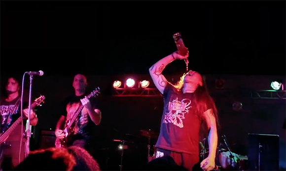 A musician in a dark t-shirt pours liquid from a bottle into his mouth on a stage with red lighting.