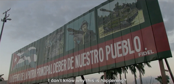 A large billboard with images of a soldier, a tank, and Spanish text reading 'LA DEFENSA DE LA PATRIA PRINCIPAL DEBER DE NUES