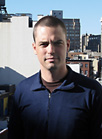 A man with short dark hair wearing a dark blue zip-up shirt against a city background.