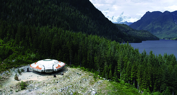 A white and orange saucer-shaped craft sits in a forest clearing overlooking a lake and mountains.