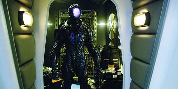 A person in dark, futuristic armored suit with a glowing purple visor stands in a doorway.