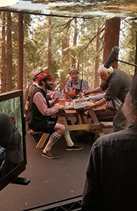 Four men sit at a wooden picnic table in front of a large forest mural during a production.