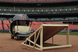 A mascot in a blue vehicle sits on a baseball field next to a large wooden ramp.