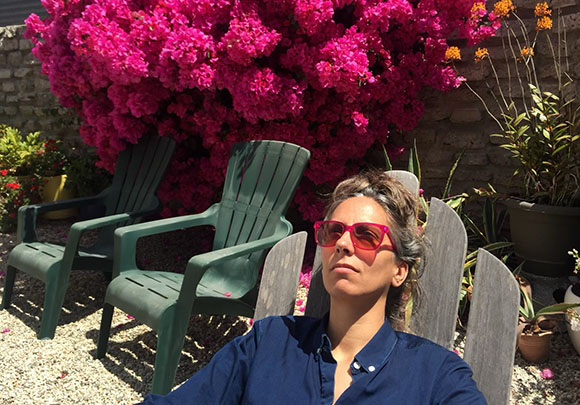 A woman in sunglasses relaxes outdoors near green chairs and large pink flowering bushes.