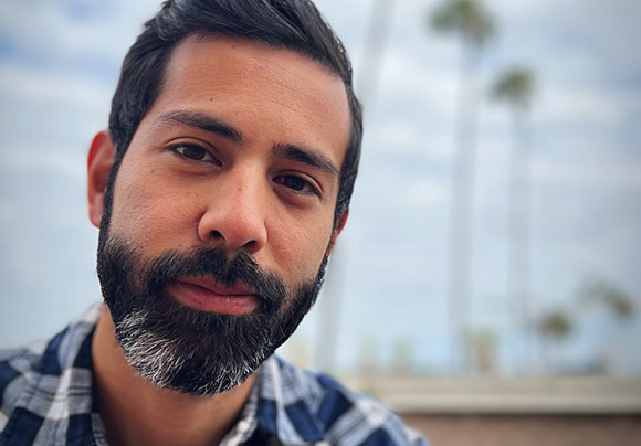 Close-up portrait of a man with dark hair and a salt-and-pepper beard against a blurred outdoor background.