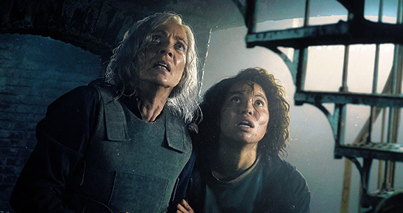 An older woman in tactical gear and a younger woman look up with fearful expressions in a dark setting.