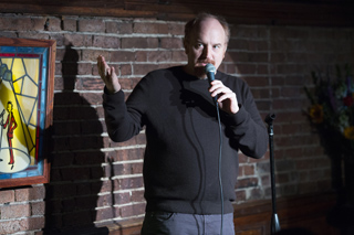 A man in a black sweater holds a microphone and gestures with his right hand against a brick wall.