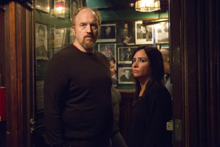 A man in a dark sweater and a woman in a black shirt stand in a room filled with framed portraits.